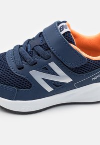 Granatowe dziecięce sneakersy z siateczkowej tkaniny, białą podeszwą, rzepem, sznurowadłami, odblaskowym logo "N" oraz pomarańczowym wnętrzem.