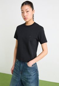Camiseta de algodón negra, de mangas cortas, con cuello redondo y diseño ajustado. Presenta un pequeño y sutil logotipo en el pecho. Combinada con jeans de mezclilla azules.