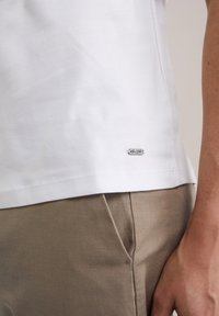 Tricou alb din bumbac cu croi drept, dotat cu un mic etichete de logo argintie la tiv, asociat cu pantaloni din stofă bej deschis.