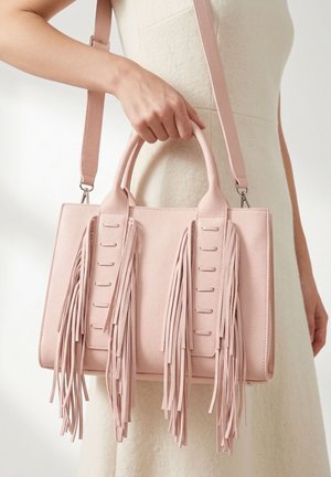 Donna che tiene una borsa a mano in pelle rosa chiaro con lunghe frange e una tracolla staccabile, su sfondo neutro.