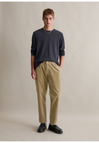 Marineblaues Langarmhemd, beige Kordhosen mit vertikalen Streifen und schwarze Schuhe. Das Modell steht mit den Händen in den Taschen vor einem neutralen Hintergrund.