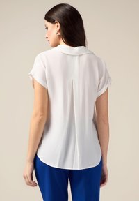 Witte blouse met korte mouwen, een kraag en een centrale rugplooi. De stof lijkt lichtgewicht en soepel, met een licht afgeronde zoom.