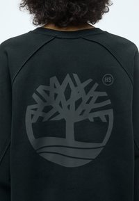 Sudadera negra con un gran logo de árbol gris texturizado y las letras "HS" en la parte trasera. La tela es suave y tiene un diseño de cuello redondo.