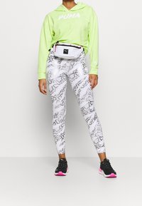 Hoodie vert néon avec logo blanc, leggings imprimé serpent blanc, et chaussures de sport noires avec des accents roses. Un sac banane blanc porté à la taille.