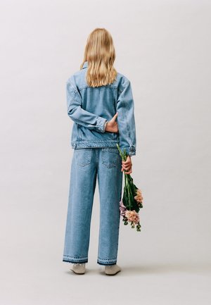 Osoba o długich blond włosach, ubrana w dżinsową kurtkę i dżinsy, stoi tyłem, trzymając mały bukiet różowych i fioletowych kwiatów.