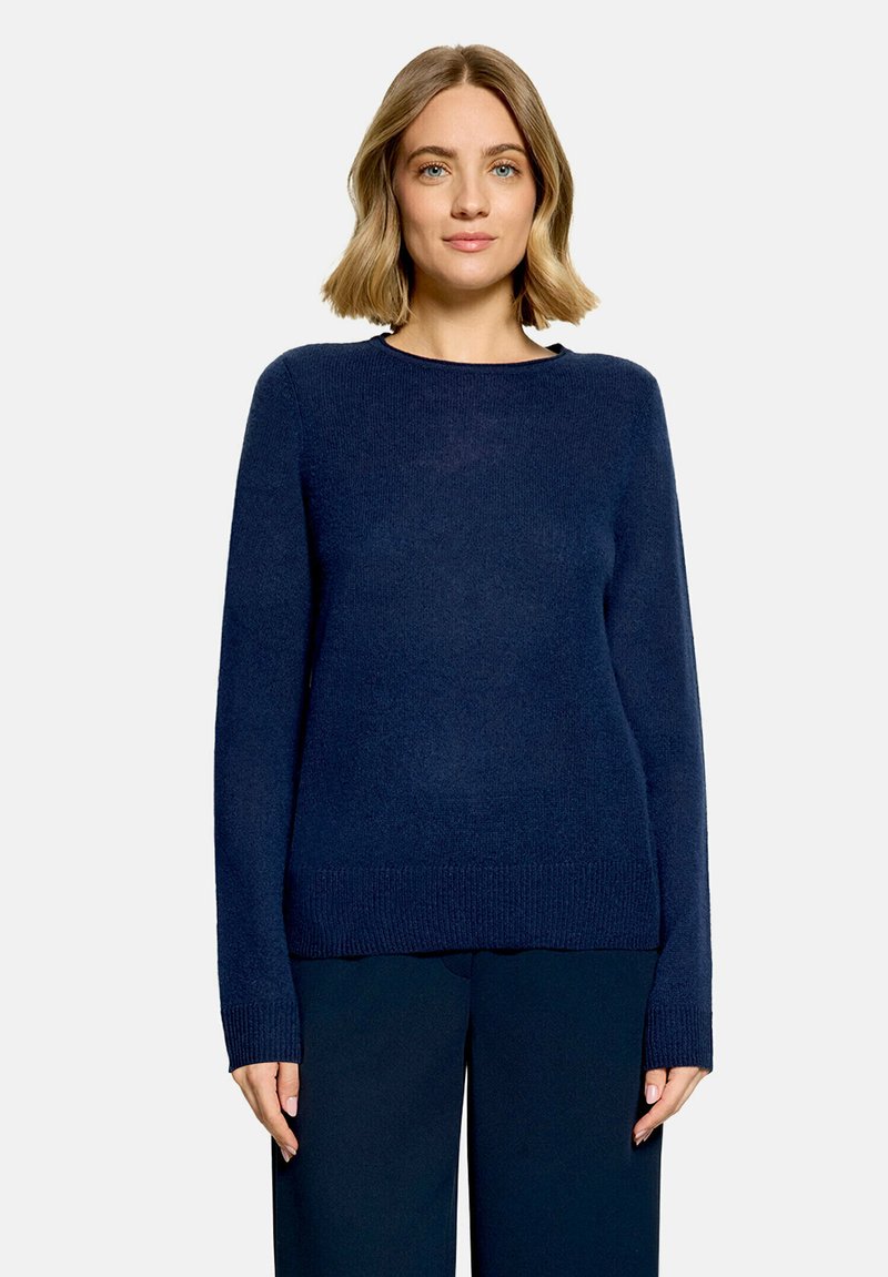 Femme aux cheveux blonds portant un pull à manches longues bleu foncé et un pantalon bleu foncé assorti, debout sur un fond clair uni.