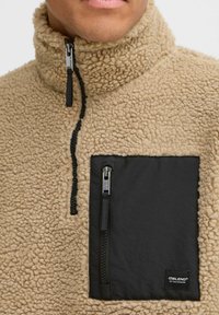 Beige fleece pullover with a high collar, featuring a black zippered chest pocket and a textured fabric finish throughout.