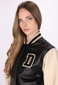 Zwarte leren varsityjack met crèpekleurige mouwen, voorzien van een opvallende geborduurde "D" en gestreepte kraagdetails.