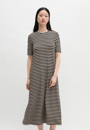Kvinde med langt sort hår iført en beige og sort midi-kjole med vandrette striber og korte ærmer, stående mod en ensfarvet lys baggrund.