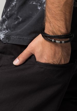 Main masculine reposant dans la poche d’un pantalon noir, portant des bracelets en cuir tressé noir avec des fermoirs en argent, sur un t-shirt noir et gris tie-dye.