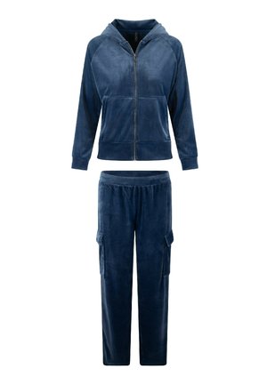 Löyhkönsininen velour-urheiluasu, jossa on vetoketjullinen huppari ja yhdistelevät cargo-housut, joissa on sivutaskut ja sileä pinta.