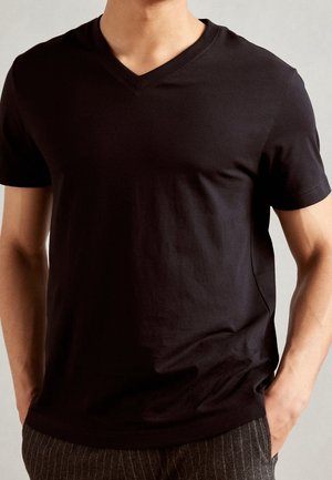 Mężczyzna ubrany w prostą czarną koszulkę z dekoltem w serek i ciemne, paskowane spodnie, z rękami w kieszeniach, na neutralnym tle.