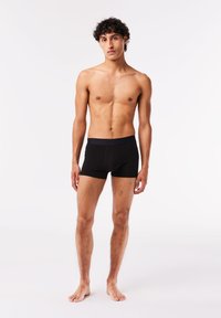 Shorts de boxe noirs avec une texture lisse et une taille élastique basse. Le modèle se tient pieds nus avec une expression neutre sur un fond uni.