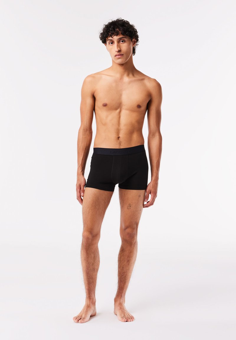 Schwarze Boxer-Shorts mit glatter Textur und einem tief sitzenden elastischen Bund. Das Modell steht barfuß mit neutralem Ausdruck vor einem einfarbigen Hintergrund.