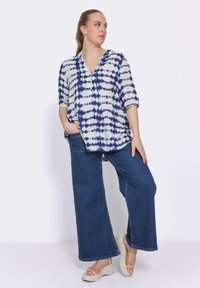 Losse blouse met korte mouwen in een marineblauw en witte tie-dye patroon, met een V-hals en knoopdetail, gecombineerd met een wide-leg spijkerbroek en platform sandalen.