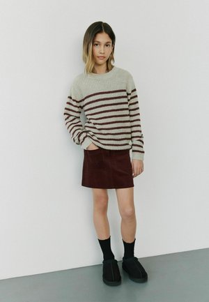 Maglione lavorato a maglia con strisce crema e bordeaux, abbinato a una gonna di velluto bordeaux. Indossa calzini neri e scarpe scure. Sfondo minimalista.