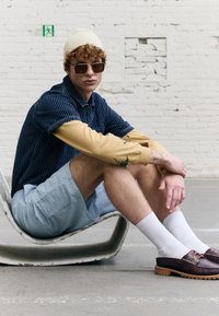 Modello seduto su una sedia scultorea, indossando una camicia a righe blu navy a maniche corte, una maglietta beige a maniche lunghe con ricami, pantaloni corti azzurri, occhiali da sole e scarpe marroni.