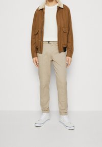 Veste marron à fermeture éclair avec un col en fausse fourrure, pull blanc texturé, pantalon beige et baskets montantes blanches sur un fond uni.