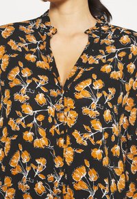 Schwarze Bluse mit orangefarbenem Blumenmuster. Verfügt über einen kragenlosen Ausschnitt und eine Knopfleiste. Leichter Stoff mit strukturierter Oberfläche.