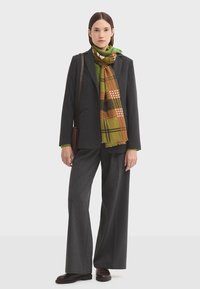 Blazer gris avec une coupe structurée, associé à un pantalon large à rayures. Écharpe verte avec un motif à carreaux aux accents orange et marron.