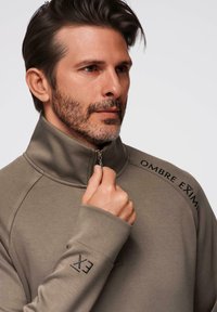 Sudadera de color marrón claro con media cremallera hecha de un tejido suave. Presenta un cuello alto, el texto negro "OMBRA EXIM" en el hombro y una cremallera prominente.