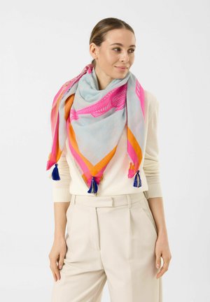 Femme portant un pull crème et un pantalon avec une grande écharpe bleu clair ornée de bordures roses et orange et de pompons bleus, regardant sur le côté