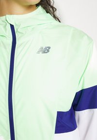 Lekka, jasnozielona kurtka z poziomym paskiem w kolorze granatowym i białym. Zapięcie na zamek błyskawiczny z odblaskowym logo New Balance.