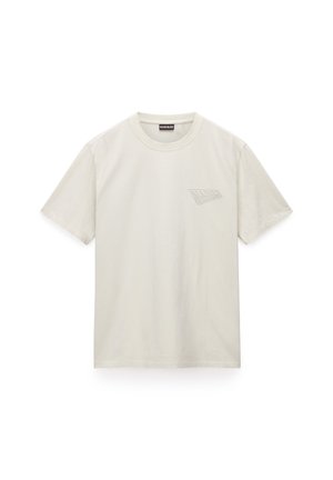 Camiseta de algodón beige claro con cuello redondo, mangas cortas y un logo impreso sutil en el lado izquierdo del pecho. Textura suave.