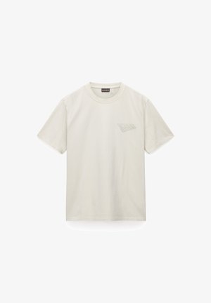 Camiseta de algodón beige claro con cuello redondo, mangas cortas y un logo impreso sutil en el lado izquierdo del pecho. Textura suave.