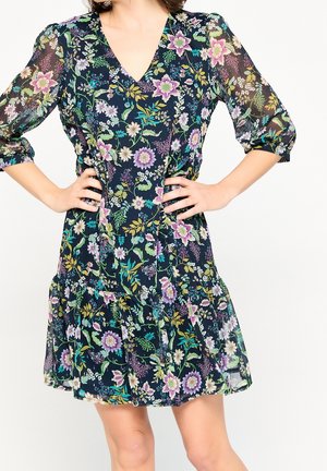 Femme en robe fleurie foncée avec manches transparentes 3/4, encolure en V et ourlet à volants, mains sur les hanches sur fond blanc uni.