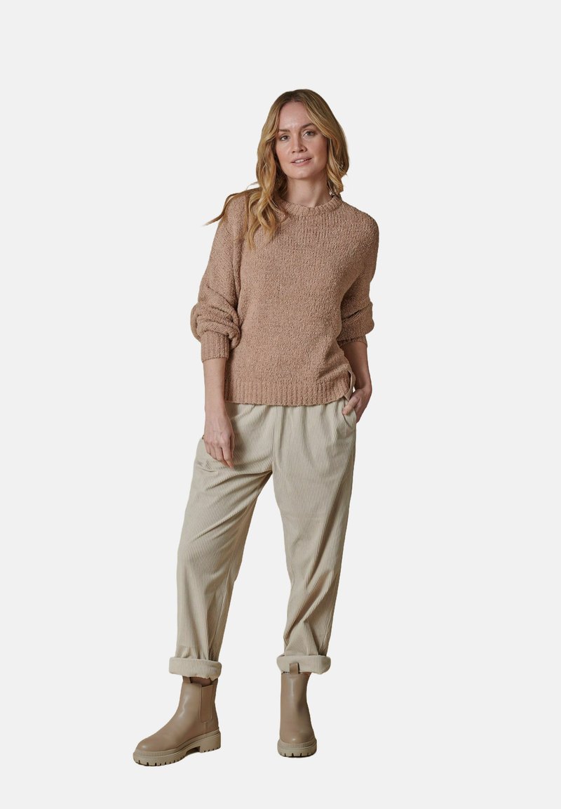 Strickpullover in Hellbraun mit umgeschlagenen Bündchen, kombiniert mit beigen, strukturierten Hosen und kastanienbraunen Stiefeletten, alles in einem lässigen Schnitt.