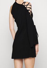 Robe noire avec un design à une épaule, détails à lacets sur la manche gauche et un accent noué à la taille. Texture lisse, longueur courte.