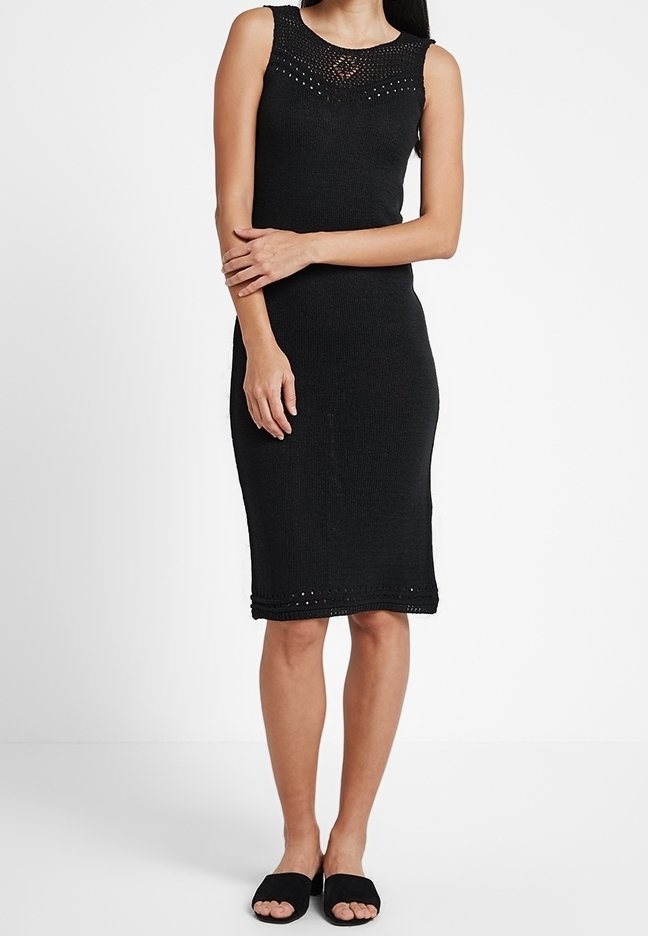 Robe tricotée noire sans manches jusqu'au genou avec un col à motif, portée avec des sandales plates noires à bout ouvert, sur une personne debout.