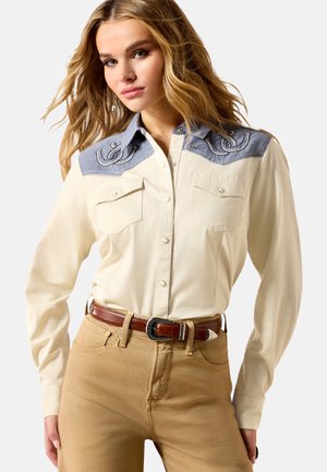 Woman wearing cream button-up shirt with denim yoke detailing and embroidered designs, tan pants, and a brown leather belt.