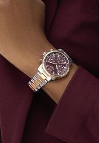 Orologio in metallo argento e oro con quadrante viola, caratterizzato da funzioni cronografiche e cinturino a motivo, indossato su un polso su uno sfondo borgogna.