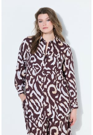 Kobieta w luźnej, dopasowanej bluzce i spodniach z abstrakcyjnym wzorem zwierzęcym w kolorach brązowym, beżowym i jasnoniebieskim, stojąca na białym tle.