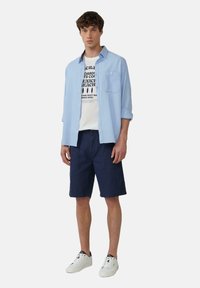 Lichtblauw overhemdd blouse gedragen over een wit T-shirt met graphics, gecombineerd met marineblauwe shorts en witte sneakers. Casual zomeroutfit.