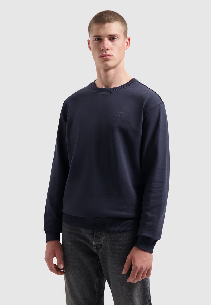 Donkerblauwe sweatshirt van zacht materiaal met rib gebreide mouwen en onderrand. Kenmerkt zich door het minimalistische "PURE PATH" logo op de borst. Klassiek ontwerp met ronde hals.