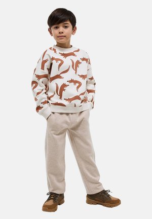 Garçon debout, les mains dans les poches, portant un sweat-shirt blanc avec des imprimés de renard marron, un pantalon beige et des chaussures de randonnée marron.