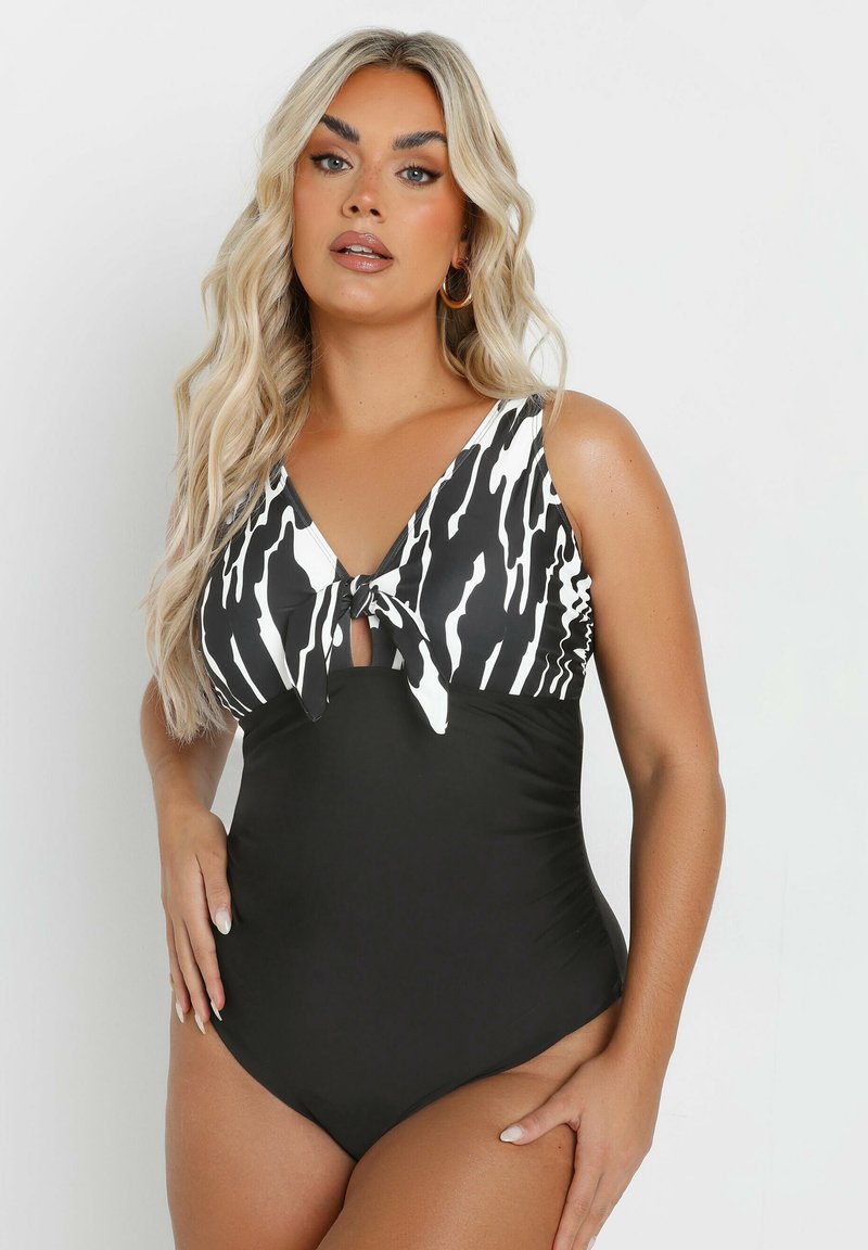 Svart baddräkt i ett plagg med ett överdel som har ett vitt och svart zebramönster, med en knutdetalj vid bröstet och ett elegant, slätt tyg.