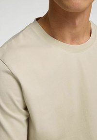 Beige cotton t-shirt with a round neckline, short sleeves, and smooth texture. Visible seams and stitching accentuate the minimalist design.