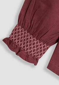 Burgundy színű blúzujj, amelyet rózsaszín, ráncolt varrás díszít a mandzsettán. Az anyag puha, kissé textúrált felületű és fodros szélű.