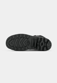 Semelle de bottes noire avec des motifs de bande de roulement profonds, comprenant des chevrons et des formes en losange, sur un fond gris clair uni.