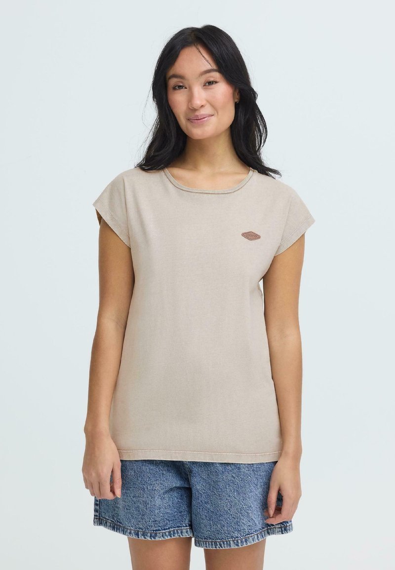 Jeune femme aux cheveux longs noirs portant un haut beige à manches courtes et un short en jean bleu, posant devant un fond clair uni.