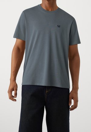 Szara bawełniana koszulka z okrągłym dekoltem, krótkimi rękawami i małym logo po lewej stronie klatki piersiowej. Projekt jest prosty i luźny.