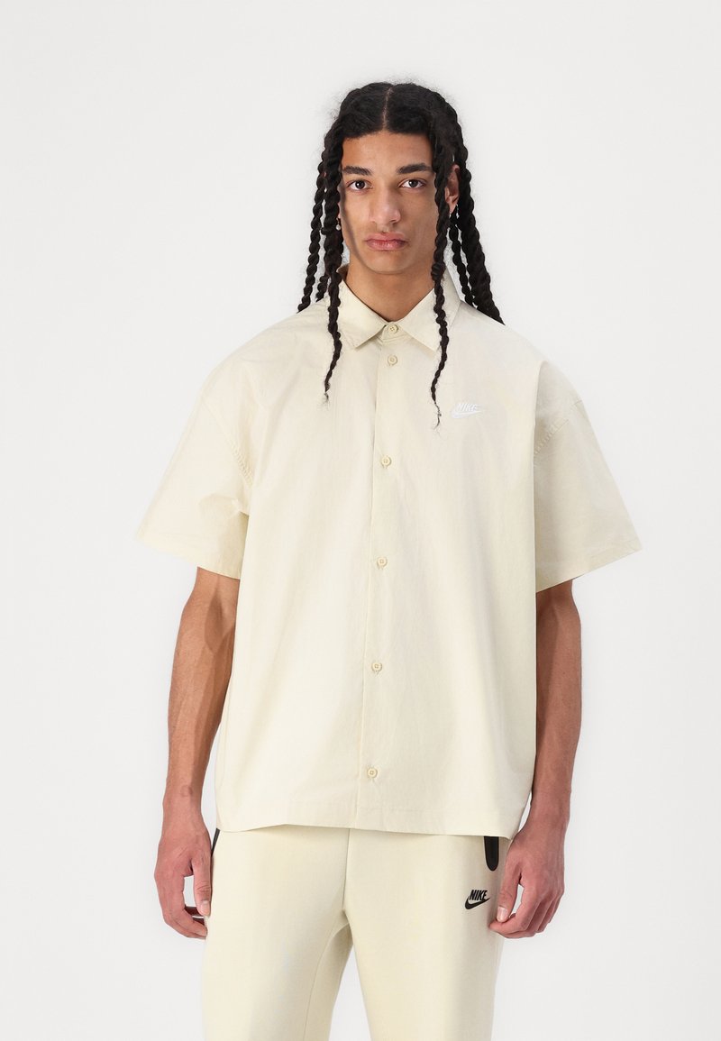 Chemise à manches courtes, beige clair, avec un col classique et un petit logo Nike blanc ; tissu en coton lisse.