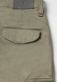 Khaki stoffen zak met afgeronde klep, gestikte randen en een rechthoekig label erboven met een reliëflogo. Glad oppervlak, geen zichtbare patronen.