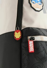 Musta ja beige reppu, jossa on Marvel-hahmojen applikaatioita; varustettu vetoketjun vedolla, joka on muotoiltu Iron Manin kypäräksi, sekä punaisella "MARVEL" -tunnisteella.