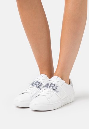 Sneakers bianche con lacci e ampie fasce in velcro decorate con testo in strass neri "KARL", indossate su gambe nude su sfondo bianco.