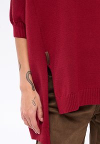 Czerwony sweter z dzianiny z prążkowanymi mankietami i dołem, z bocznym rozcięciem, metka po lewej stronie, noszony z brązowymi spodniami. Gładka faktura, luźny krój.